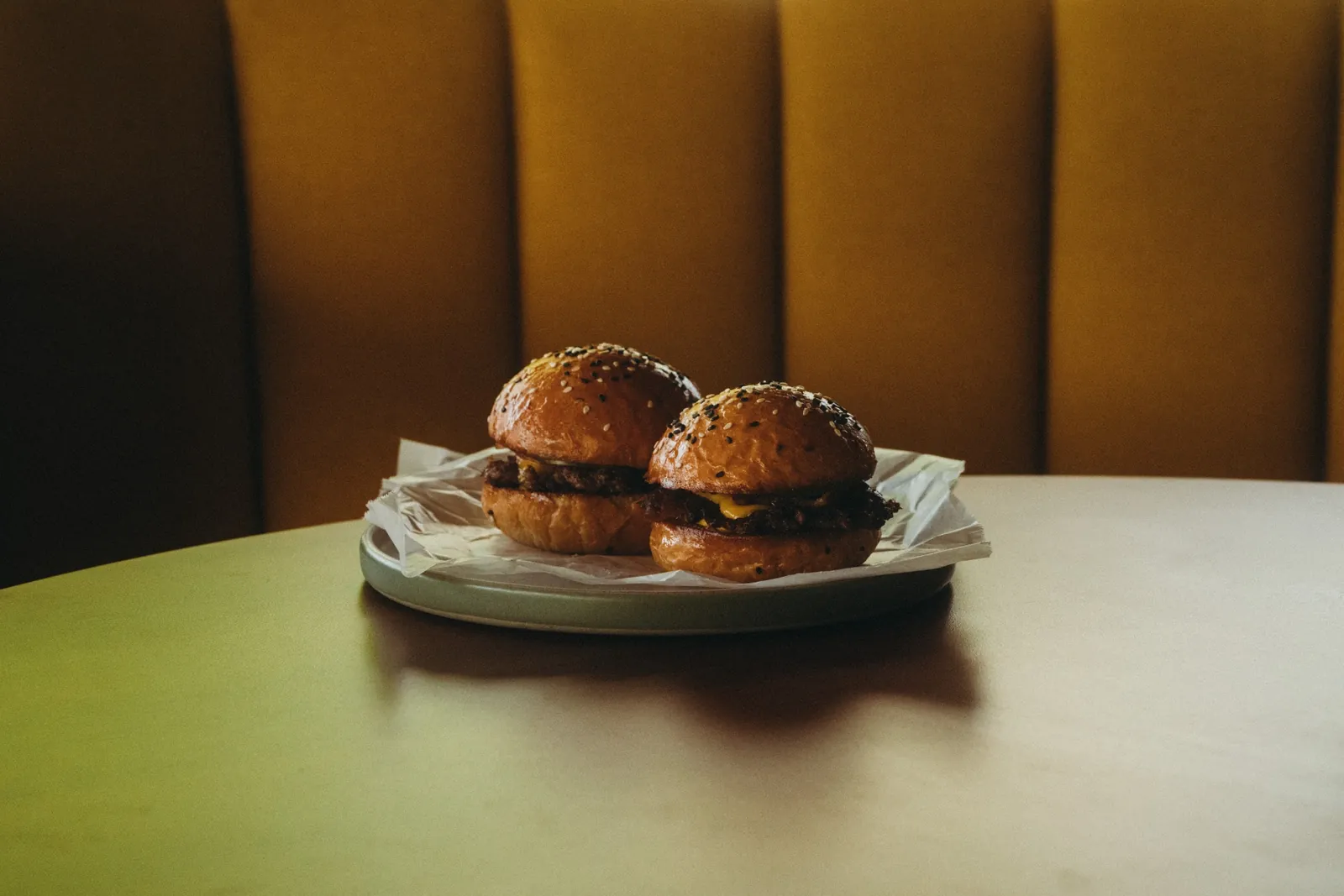 Two golden brioche cheeseburgers on a plate against a mustard velvet booth at The Lakehouse
